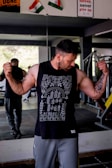 A man in a gym flexes his muscles while wearing a sleeveless black shirt with intricate white patterns. Two flags are seen in the background above a motivational poster. The gym layout is visible, including a mirror reflecting some of the interior.