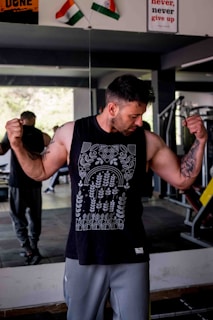 A man in a gym flexes his muscles while wearing a sleeveless black shirt with intricate white patterns. Two flags are seen in the background above a motivational poster. The gym layout is visible, including a mirror reflecting some of the interior.