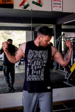 A man in a gym flexes his muscles while wearing a sleeveless black shirt with intricate white patterns. Two flags are seen in the background above a motivational poster. The gym layout is visible, including a mirror reflecting some of the interior.