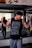 A man in a gym flexes his muscles while wearing a sleeveless black shirt with intricate white patterns. Two flags are seen in the background above a motivational poster. The gym layout is visible, including a mirror reflecting some of the interior.