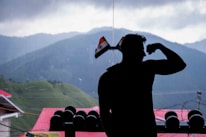 A silhouetted person is flexing their arm in front of an overcast mountainous landscape. Indian flags adorn the window beside them, and a row of dumbbells is visible. The background features lush green hills dotted with trees and buildings with red roofs.