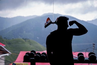 A silhouetted person is flexing their arm in front of an overcast mountainous landscape. Indian flags adorn the window beside them, and a row of dumbbells is visible. The background features lush green hills dotted with trees and buildings with red roofs.