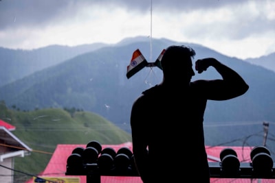 A silhouetted person is flexing their arm in front of an overcast mountainous landscape. Indian flags adorn the window beside them, and a row of dumbbells is visible. The background features lush green hills dotted with trees and buildings with red roofs.