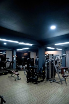 A modern gym setting with various exercise equipment such as treadmills, benches, and weight machines. The room is dimly lit with overhead fluorescent lights and features a wooden floor.