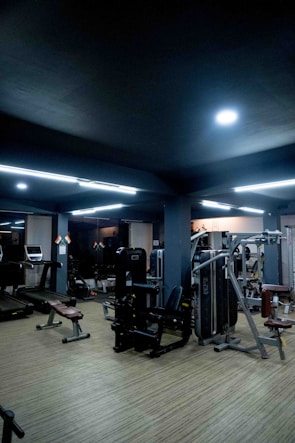 A modern gym setting with various exercise equipment such as treadmills, benches, and weight machines. The room is dimly lit with overhead fluorescent lights and features a wooden floor.