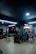 A modern gym setting with various exercise equipment such as treadmills, benches, and weight machines. The room is dimly lit with overhead fluorescent lights and features a wooden floor.