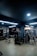 A modern gym setting with various exercise equipment such as treadmills, benches, and weight machines. The room is dimly lit with overhead fluorescent lights and features a wooden floor.
