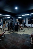 A dimly lit gym interior featuring various exercise machines and benches. The gym equipment includes weight machines and benches, arranged on a floor with dark rubber tiles. Overhead lighting provides an even illumination across the space.