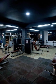 A dimly lit gym interior featuring various exercise machines and benches. The gym equipment includes weight machines and benches, arranged on a floor with dark rubber tiles. Overhead lighting provides an even illumination across the space.