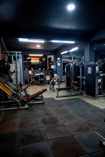A dimly lit gym with various exercise machines, including leg presses and cable equipment. The floor is padded with dark tiles. In the background, two people are sitting at a desk with a computer. An Indian flag is visible on the wall, adding a touch of decoration.