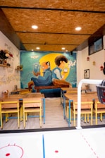 A cozy café with wooden tables and chairs arranged neatly. The back wall features a large, colorful mural depicting two cartoon-like characters, one reading a book and the other drinking a beverage, sitting back-to-back. The ceiling is made of wooden panels with recessed lighting. Decor includes small shelves with potted plants and various wall hangings, including small colorful lanterns.