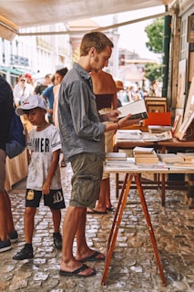 A man is standing at an outdoor market, reading a book from a table filled with various books. He's casually dressed in a gray shirt and shorts, wearing flip-flops. Next to him, a young child in a white cap, t-shirt, and shorts stands looking around. The background features other people browsing and a canopy providing shade over the cobblestone street.