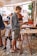 A man is standing at an outdoor market, reading a book from a table filled with various books. He's casually dressed in a gray shirt and shorts, wearing flip-flops. Next to him, a young child in a white cap, t-shirt, and shorts stands looking around. The background features other people browsing and a canopy providing shade over the cobblestone street.