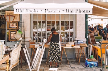 A woman stands in front of a vintage shop with various items like old books, posters, and photos displayed on tables. The shop has a quaint and rustic appearance, with framed pictures and antique furniture visible. Several people are nearby, possibly browsing or interacting.