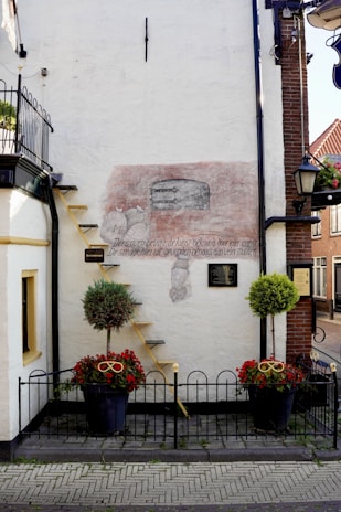 A white building wall featuring a faded mural with a simple sketch and some text in another language. The mural includes imagery of two figures and an oven. A small yellow ladder leans against the wall, reaching up to a first-story balcony protected by a black wrought iron railing. Below, the base of the wall has two large pots containing green shrubs and red flowers. A black metal fence encloses the plant area.