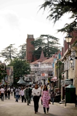 A bustling street scene with numerous people walking and interacting in a charming town setting. The architecture features a mix of traditional and modern building styles, with timber-framed facades and colorful details. The street is lined with trees, and a prominent tower rises in the background amidst tall green trees. The atmosphere suggests a lively and busy environment typical of a tourist or local city center.