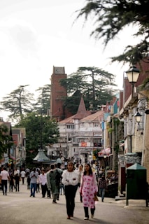 A bustling street scene with numerous people walking and interacting in a charming town setting. The architecture features a mix of traditional and modern building styles, with timber-framed facades and colorful details. The street is lined with trees, and a prominent tower rises in the background amidst tall green trees. The atmosphere suggests a lively and busy environment typical of a tourist or local city center.