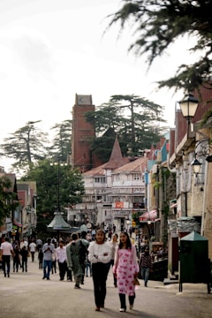 A bustling street scene with numerous people walking and interacting in a charming town setting. The architecture features a mix of traditional and modern building styles, with timber-framed facades and colorful details. The street is lined with trees, and a prominent tower rises in the background amidst tall green trees. The atmosphere suggests a lively and busy environment typical of a tourist or local city center.