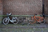 Three bicycles are parked against a brick wall. One of the bicycles is black and appears to be in a damaged condition with the frame lying on the ground. The other two are upright, one being white with a baby seat attached, and the other orange. The ground consists of cobblestone pavement.