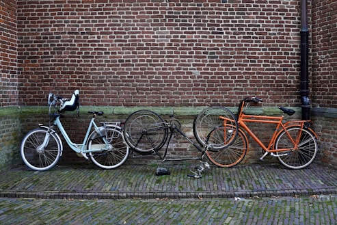Three bicycles are parked against a brick wall. One of the bicycles is black and appears to be in a damaged condition with the frame lying on the ground. The other two are upright, one being white with a baby seat attached, and the other orange. The ground consists of cobblestone pavement.