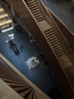 An indoor architectural space is viewed from above, featuring multiple wooden staircases and a spacious open area. People are seen walking and interacting on different levels, and there is a white tent-like structure in the center of the floor. The lighting creates a pattern of shadows on the ground.