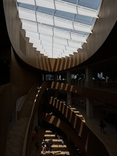 A modern interior space featuring a large skylight with geometric patterns casting shadows across multiple levels of a public area. The structure is made primarily of wood, creating a warm and inviting atmosphere. People are casually walking and sitting on benches, giving the space a communal feel.