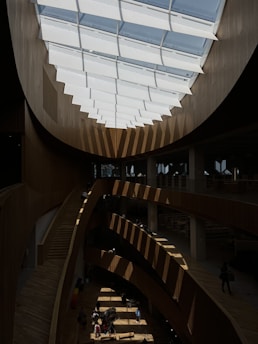 A modern interior space featuring a large skylight with geometric patterns casting shadows across multiple levels of a public area. The structure is made primarily of wood, creating a warm and inviting atmosphere. People are casually walking and sitting on benches, giving the space a communal feel.