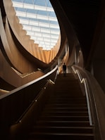 An interior architectural scene featuring a grand wooden staircase leading upwards, illuminated by natural light from a large, segmented skylight. The design elements include sweeping, curved wooden panels and railings with integrated lighting. A solitary person is seen ascending the stairs, adding a sense of scale to the space.