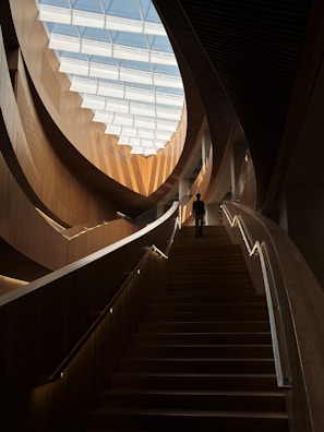 An interior architectural scene featuring a grand wooden staircase leading upwards, illuminated by natural light from a large, segmented skylight. The design elements include sweeping, curved wooden panels and railings with integrated lighting. A solitary person is seen ascending the stairs, adding a sense of scale to the space.