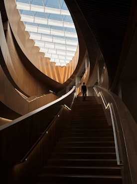 An interior architectural scene featuring a grand wooden staircase leading upwards, illuminated by natural light from a large, segmented skylight. The design elements include sweeping, curved wooden panels and railings with integrated lighting. A solitary person is seen ascending the stairs, adding a sense of scale to the space.