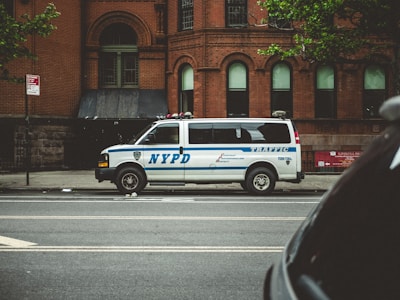 A white NYPD traffic enforcement van is parked on the side of a city street in front of a historic brick building. The van is marked with blue stripes and several official logos, and there is a red traffic sign posted nearby. Green leafy branches partially extend into view from the top right corner.