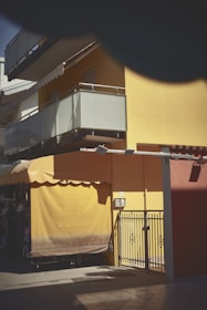 A yellow and red building with a covered balcony and awnings. The bottom floor has a closed yellow awning and a metal gate. Shadows from a nearby object partially obscure the view.