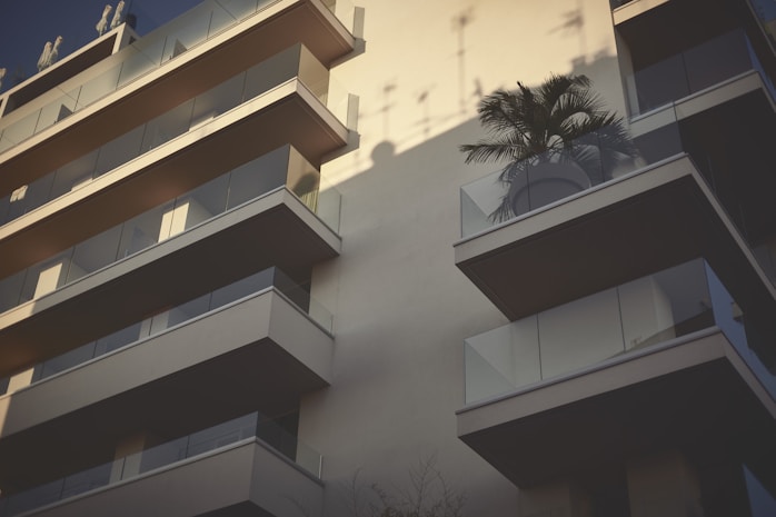 A modern apartment building featuring multiple balconies with glass railings. One balcony displays a large potted palm tree, casting intricate shadows on the wall behind. The angle of the shot highlights the architectural design, emphasizing clean lines and minimalistic elements.