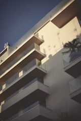 A modern apartment building featuring multiple balconies. The structure is bathed in soft, late-afternoon light, creating distinct shadows against the ivory facade. A potted palm is visible on one of the balconies, adding a tropical touch.