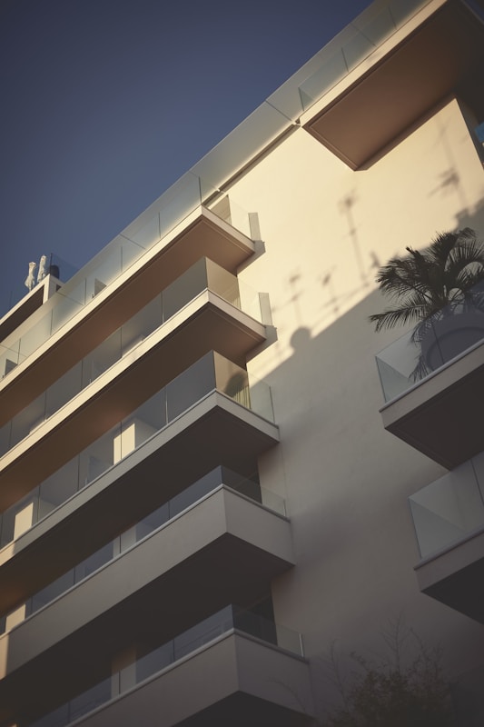 A modern apartment building featuring multiple balconies. The structure is bathed in soft, late-afternoon light, creating distinct shadows against the ivory facade. A potted palm is visible on one of the balconies, adding a tropical touch.