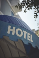 A large blue awning with the word 'HOTEL' prominently displayed in bold, white capital letters. Above the awning, parts of a building with balconies and some foliage from a tree are visible.