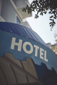 A large blue awning with the word 'HOTEL' prominently displayed in bold, white capital letters. Above the awning, parts of a building with balconies and some foliage from a tree are visible.