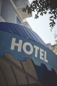 A large blue awning with the word 'HOTEL' prominently displayed in bold, white capital letters. Above the awning, parts of a building with balconies and some foliage from a tree are visible.