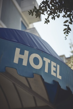 A large blue awning with the word 'HOTEL' prominently displayed in bold, white capital letters. Above the awning, parts of a building with balconies and some foliage from a tree are visible.