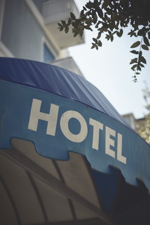 A large blue awning with the word 'HOTEL' prominently displayed in bold, white capital letters. Above the awning, parts of a building with balconies and some foliage from a tree are visible.