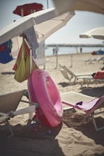A beach scene featuring several beach chairs and umbrellas. A bright pink inflatable swim ring is leaning against one of the chairs. There are various beach bags and clothes hanging from the umbrella. In the background, the sandy beach extends to the water, where a few people are visible near the shoreline. The overall atmosphere appears calm and sunny.