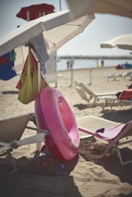 A beach scene featuring several beach chairs and umbrellas. A bright pink inflatable swim ring is leaning against one of the chairs. There are various beach bags and clothes hanging from the umbrella. In the background, the sandy beach extends to the water, where a few people are visible near the shoreline. The overall atmosphere appears calm and sunny.
