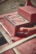 A close-up view of a red lifeguard rescue boat with the number 32 on a horizontal surface. A life buoy is attached to it with a white rope, and an oar or paddle is fastened alongside. The background shows a sandy beach with slightly blurred footwear.