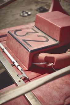 A close-up view of a red lifeguard rescue boat with the number 32 on a horizontal surface. A life buoy is attached to it with a white rope, and an oar or paddle is fastened alongside. The background shows a sandy beach with slightly blurred footwear.