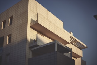 A modern building with a minimalist design, featuring a geometric facade made of light-colored tiles. The structure includes large rectangular windows and balconies with glass railings. The shadow play on the surface adds depth and contrast to the scene, with a touch of greenery visible on one of the balconies.
