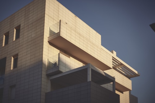 A modern building with a minimalist design, featuring a geometric facade made of light-colored tiles. The structure includes large rectangular windows and balconies with glass railings. The shadow play on the surface adds depth and contrast to the scene, with a touch of greenery visible on one of the balconies.
