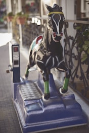 A coin-operated mechanical horse ride is positioned on a sidewalk, featuring a black and glossy finish with green accents on its legs and a decorative yellow band around its head. The ride is mounted on a blue base, adjacent to a rail decorated with intricate metalwork. In the background, blurred potted plants add to the atmosphere of an outdoor setting.