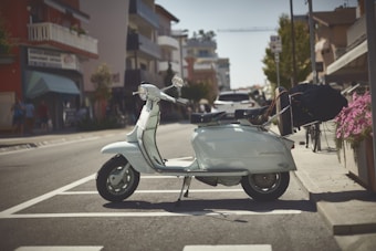 A vintage scooter is parked on a sunlit city street, positioned within a parking space lined with white stripes. The background features urban architecture with modern buildings, as well as a few street elements like flowers and parked cars. The scene has a leisurely, everyday setting with some blurred figures walking in the distance.