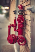A red fire hydrant system is mounted on a brick wall. The hydrant has multiple valves and connectors, with one large valve in the foreground and a pipe extending upwards.