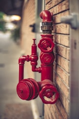 A red fire hydrant system is mounted on a brick wall. The hydrant has multiple valves and connectors, with one large valve in the foreground and a pipe extending upwards.